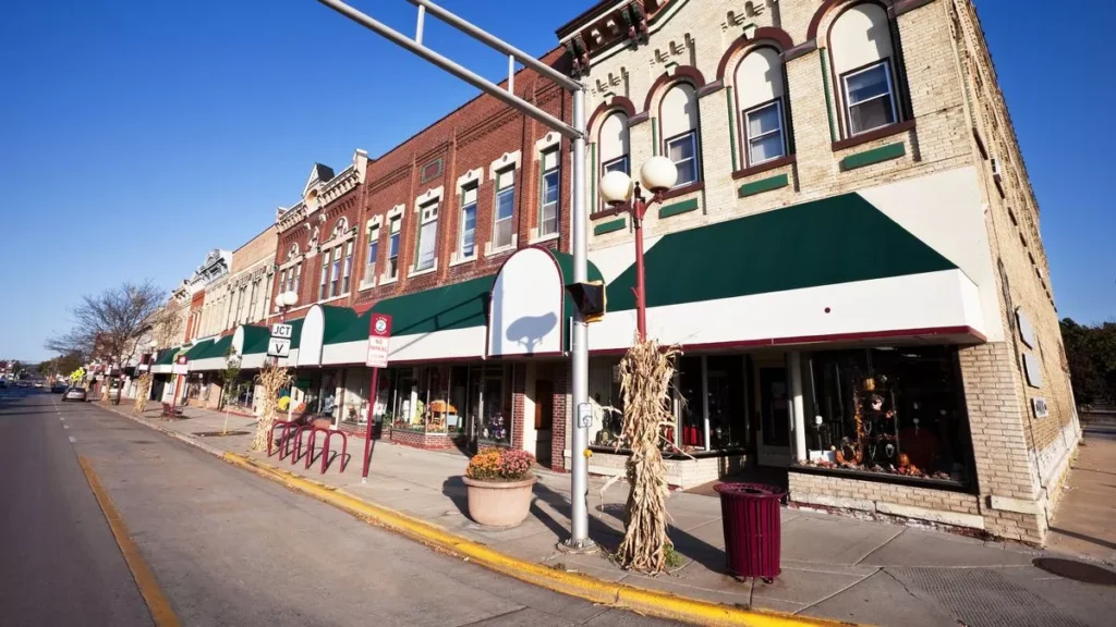 A street view of a town in Reedsburg, Wisconsin, featuring several old brick buildings with storefronts along a sidewalk. The sky is blue and clear.