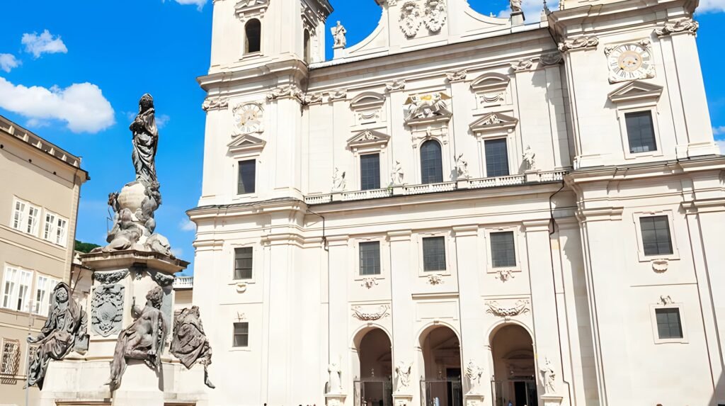 Public squares: Domplatz in Salzburg, Austria, featuring the baroque Salzburg Cathedral façade and Marian statue against a clear blue sky.