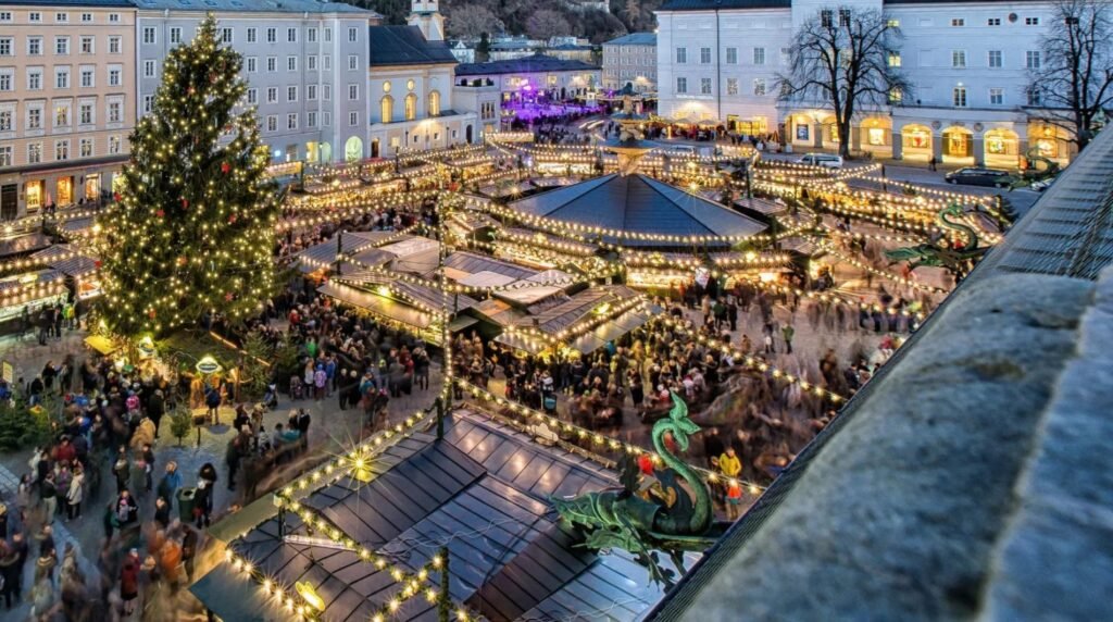 Overhead view of Krampuslauf Salzburg in Domplatz at dusk, with market stalls, a towering lit Christmas tree, and throngs of bundled spectators.