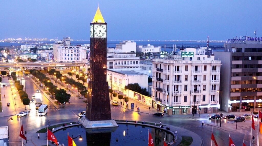 Republic Square in Tunis, Tunisia, featuring the illuminated central clock tower, surrounding buildings, and Tunisian flags at dusk.