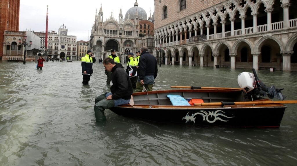 Acqua Alta Flood 2019: police and residents wade through knee-deep water beside a small boat in Piazza San Marco, with St Mark’s Basilica and the Doge’s Palace behind.