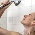 A person with light skin and light-colored hair rinses shampoo from their hair with a handheld shower head, illustrating Showering Habits.