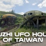 Two abandoned, UFO-shaped pod houses from the Sanzhi UFO Houses project in Taiwan sit elevated on concrete stilts amidst overgrown vegetation and a sloping road, reflecting the site's futuristic yet decayed architecture.
