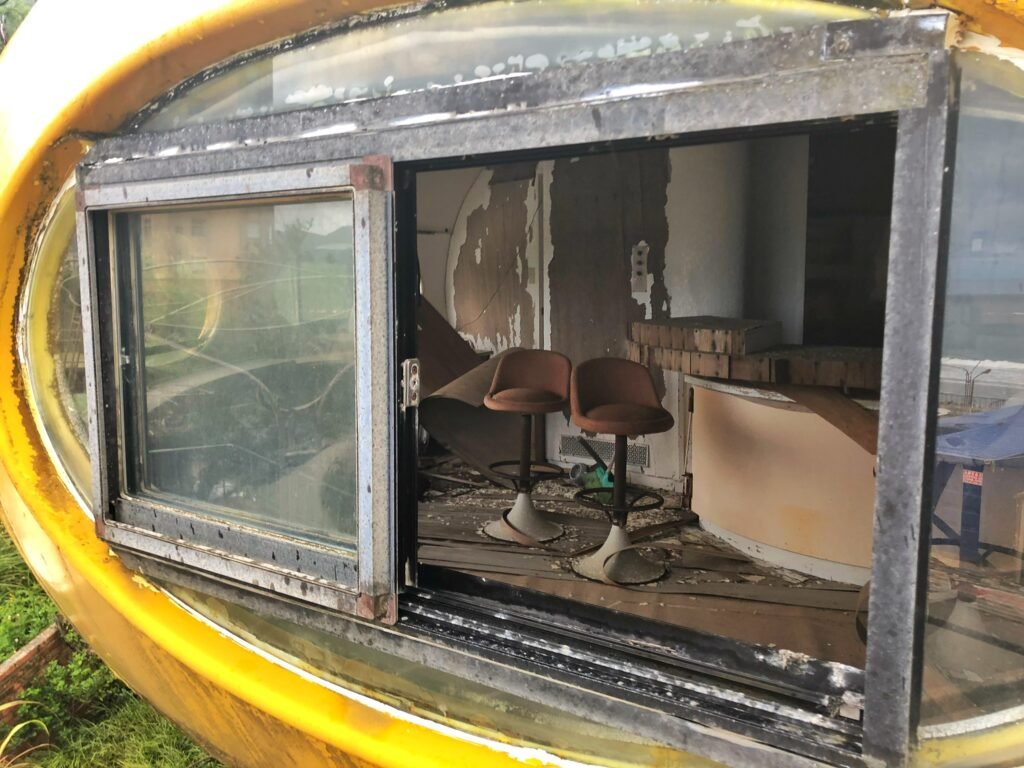 Interior view of a yellow pod showing decayed furniture and peeling walls through a broken window.