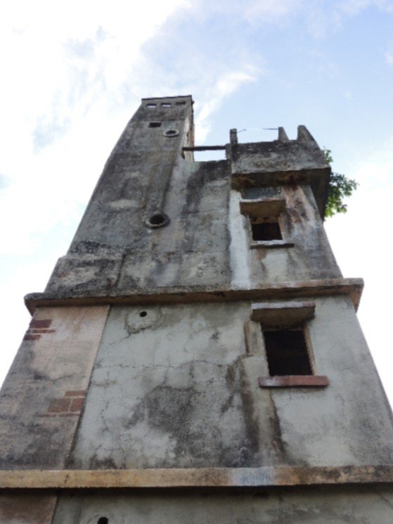 A low-angle view of a weathered concrete tower, with small windows and an unfinished top section, reaching towards a cloudy sky.