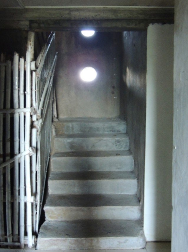 A narrow, dimly lit concrete staircase leads upwards, illuminated by two bright circular openings in the wall, with a bamboo-barred gate visible on the left.
