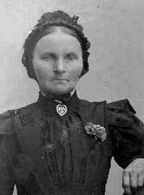 A sepia-toned bust portrait of Ane Andersdatter, a woman with a stern expression, wearing a dark dress with a high collar and a dark, ornate bonnet.