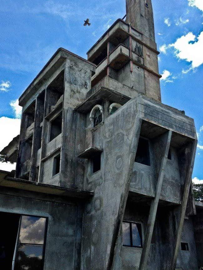 A unique, multi-level concrete structure, the Tower of Bobby Capiz, with a distinct spire and varied window designs, stands tall against a vibrant blue sky with scattered clouds.