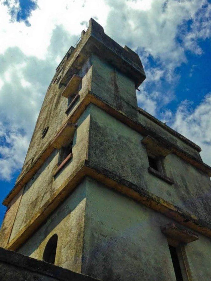 A low-angle view of a weathered concrete tower, with multiple levels and small windows, reaching dramatically towards a partly cloudy blue sky.