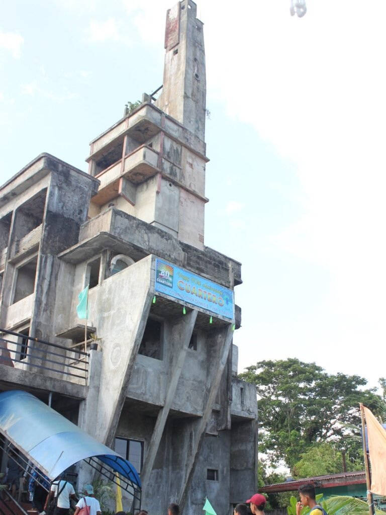 A towering, seven-story concrete structure, the Tower of Bobby Capiz, stands as a resilient landmark with unique angles and an unfinished spire against a clear sky.
