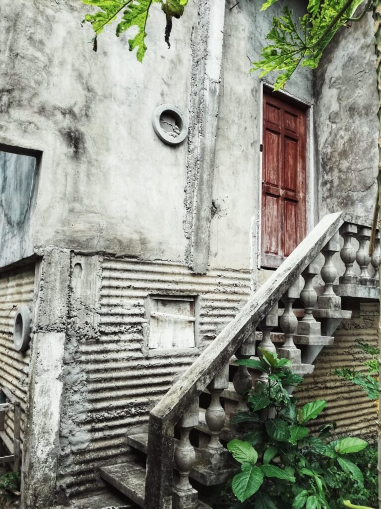 A worn, textured concrete wall with unique corrugated patterns and a small circular window, featuring an aged wooden door and a stone staircase with an ornate balustrade leading upwards.