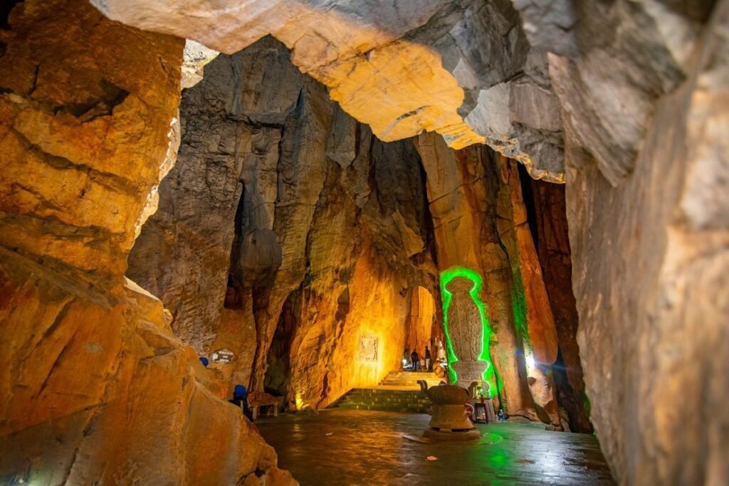 A wide view inside a cave with tall rock walls lit in warm yellow and orange. A central, slender statue or formation glows vibrantly green, with people visible at its base.
