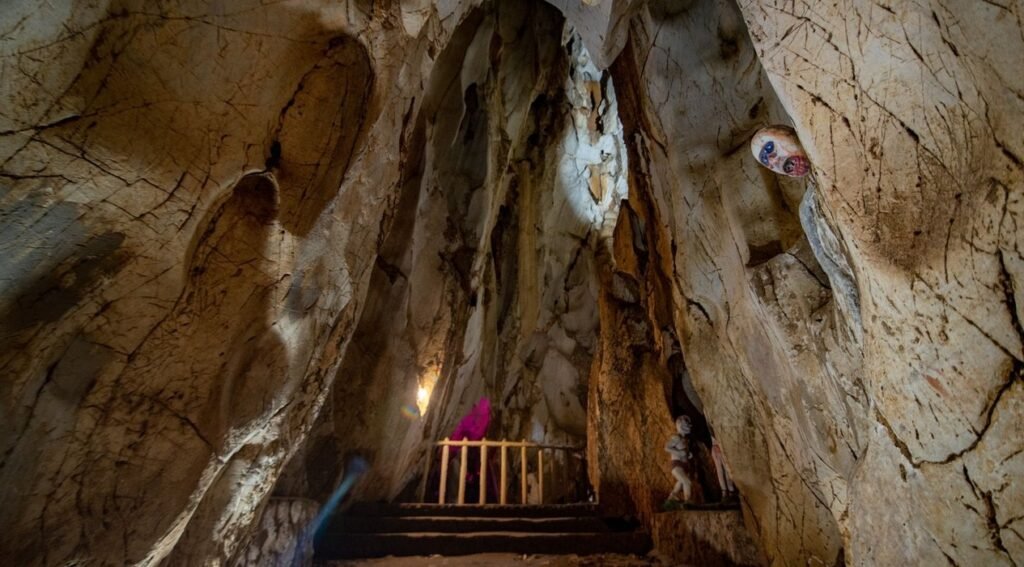 A wide-angle shot of the inside of a large cave with light brown, textured rock walls. In the center, a set of steps leads up to a small, dark area partially obscured by a pink fabric and a wooden railing. To the right of the steps, a statue of a figure with light skin and dark hair stands against the cave wall. Higher up on the right wall, a colorful, round object with what appears to be an eye design is visible in an alcove.