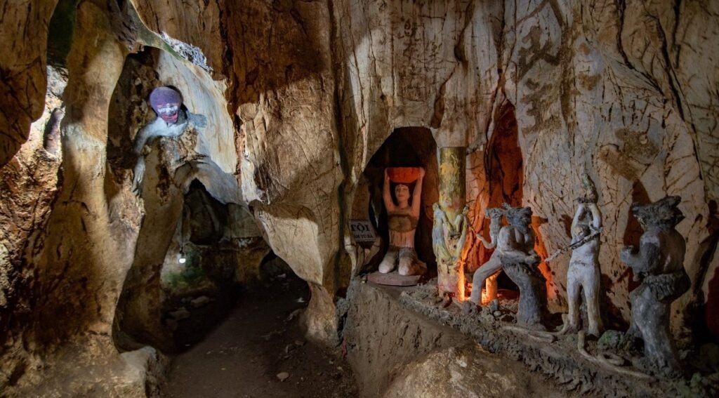 Inside a cave, several light-colored statues depict figures in distress. One figure with a purple head is on the left, while others are in an alcove on the right.
