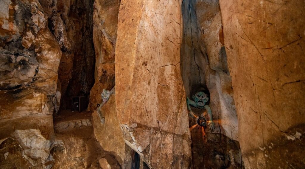 Inside a cave, a green-skinned, dark-haired figure with an angry expression and a glowing orb on its chest is nestled between two large rock formations.