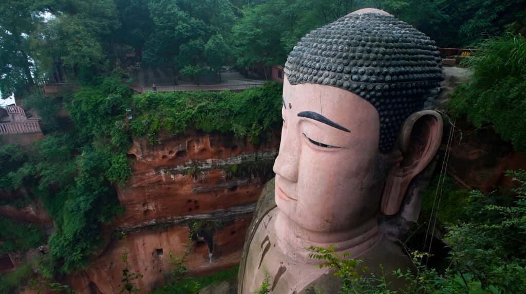A detailed side view of the Leshan Giant Buddha’s massive stone head carved into a red sandstone cliff, surrounded by lush green trees and natural rock formations, showcasing the statue’s serene expression and impressive scale.