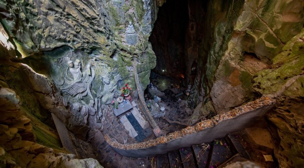 Looking down into a cave, a winding staircase with a stone railing descends into darkness. Intricate carvings of figures are etched into the left cave wall.