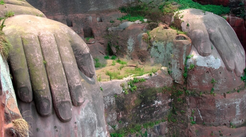 A close-up of the massive stone toes of the Leshan Giant Buddha, showing weathering effects including moss and small plants growing on the surface, illustrating the statue’s exposure to natural elements over many centuries.