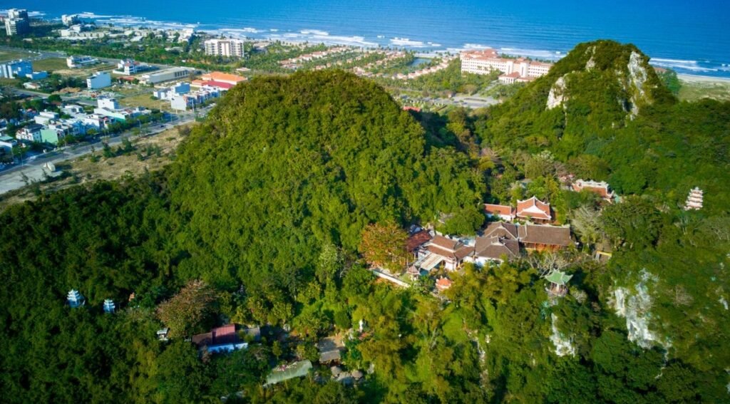 Lush green Marble Mountains in Da Nang, Vietnam, with traditional temples nestled amongst the trees and a glimpse of the coastline. Am Phu Cave is located within this sacred complex.