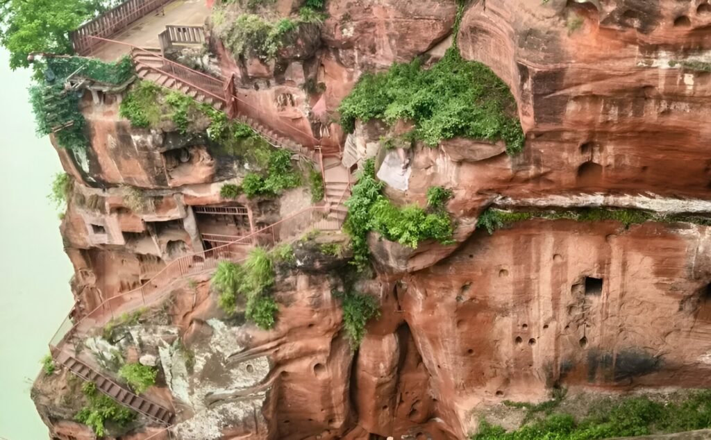 A side view of the red sandstone cliff carved with winding staircases leading up to the Leshan Giant Buddha, surrounded by lush green vegetation, showcasing the blend of natural landscape and human engineering that allows visitors to ascend and explore the statue up close.
