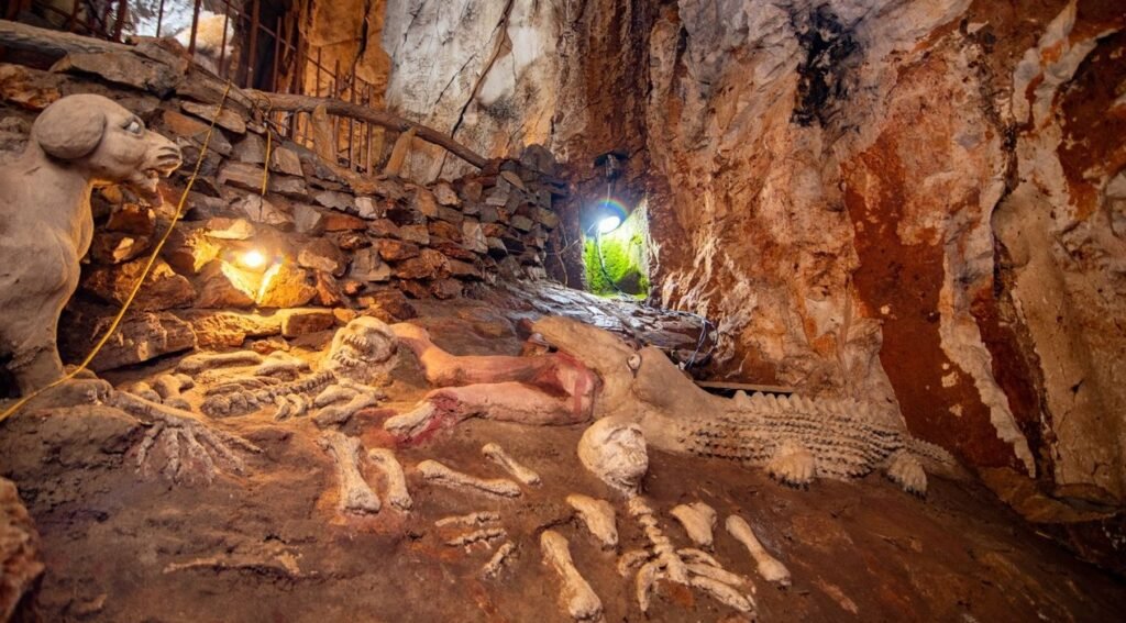 Inside a cave, a large, light-colored dog statue stands guard over skeletal remains and a decomposing crocodile-like figure on the ground. A bright opening is visible ahead.