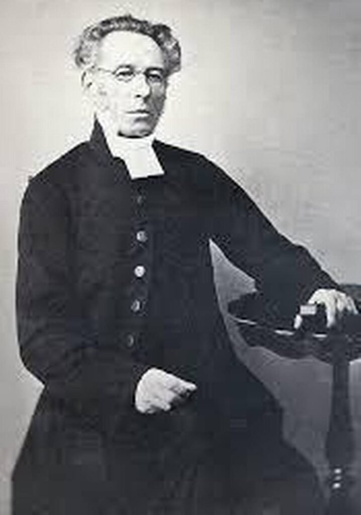 A vintage black and white portrait of Anders Lindbäck, a man with glasses and curly hair, wearing a dark clerical coat and a white collar, seated with one hand resting on a table.