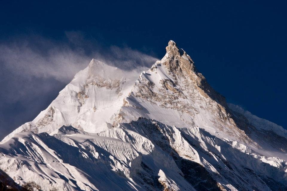 Manaslu, the eighth highest mountain on Earth at 8,163 meters, showing sharp rocky and snowy peaks with wind blowing snow, located in Nepal under a clear blue sky.