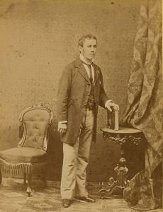 A sepia-toned full-body portrait of Ferdinand Wittmann, a young man in a suit, standing beside a small table with a book, with a patterned chair and draped fabric behind him.