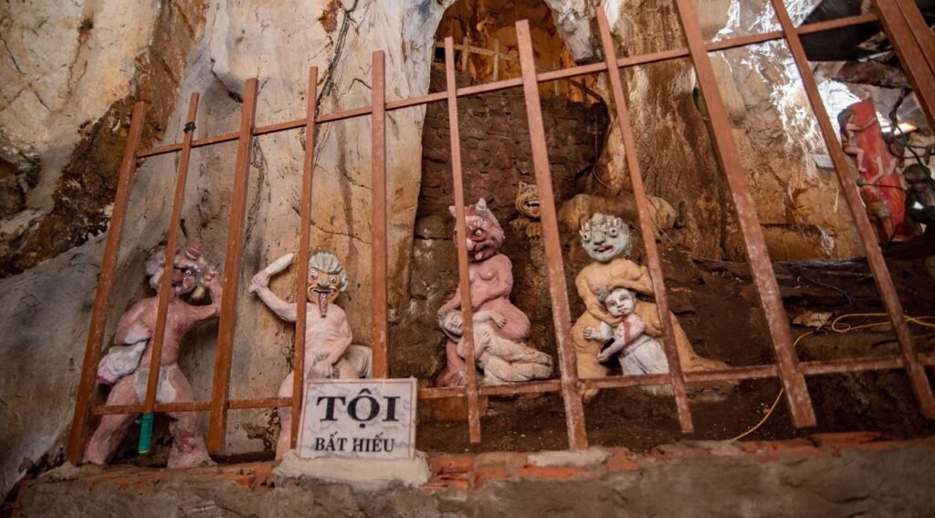 Inside a cave, several grotesque, light-colored statues depict tortured figures, some with distorted faces and limbs. They are positioned behind a wooden bar structure.