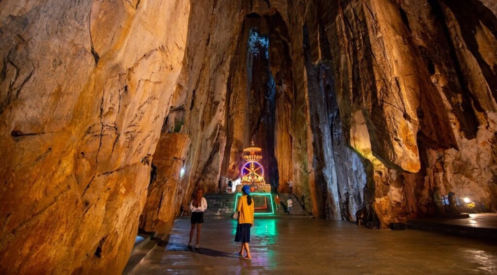 Inside a vast cave, a brightly lit shrine with a wheel-like design glows purple and green in the distance. Two visitors stand on the damp cave floor in the foreground.