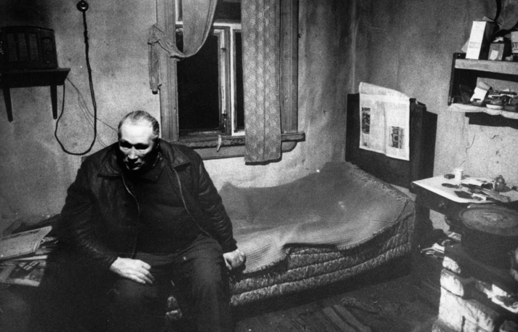 A grim, black and white photograph of Pentti Saarinen sitting on a mattress in a sparsely furnished, dark room, years after he was suspected of murdering his wife.