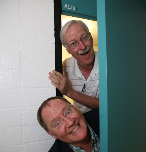 Pixar legends John Lasseter and another colleague smiling at the CalArts classroom door marked “A113,” the origin of the Easter egg.