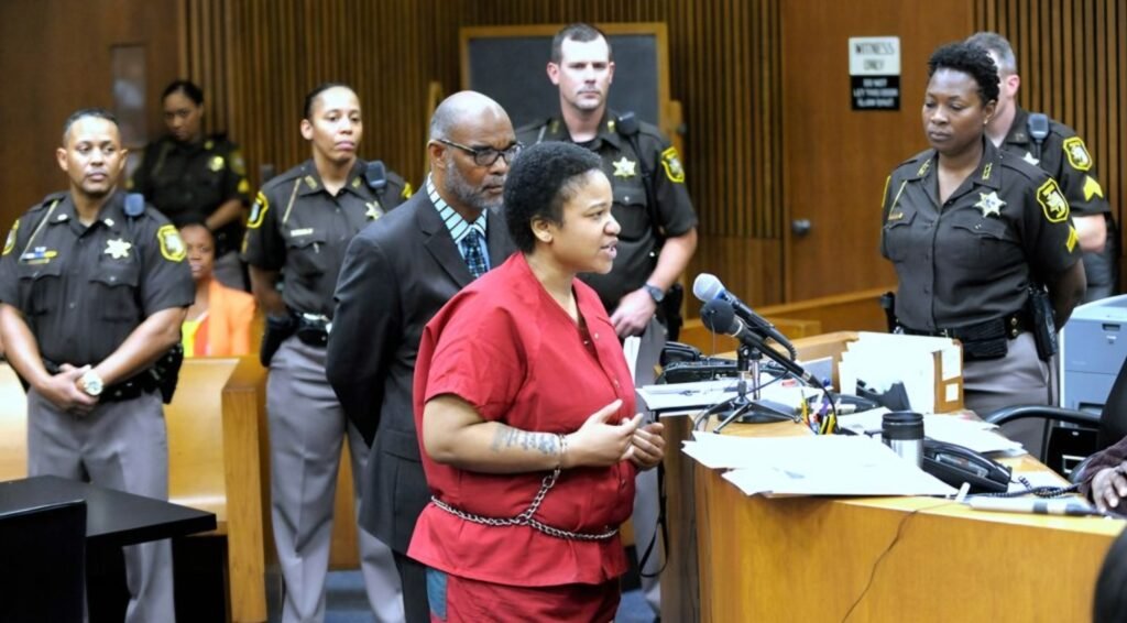 Mitchelle Blair, in a red jumpsuit, addresses the court from a podium, surrounded by her lawyer and multiple sheriff's deputies during her trial.